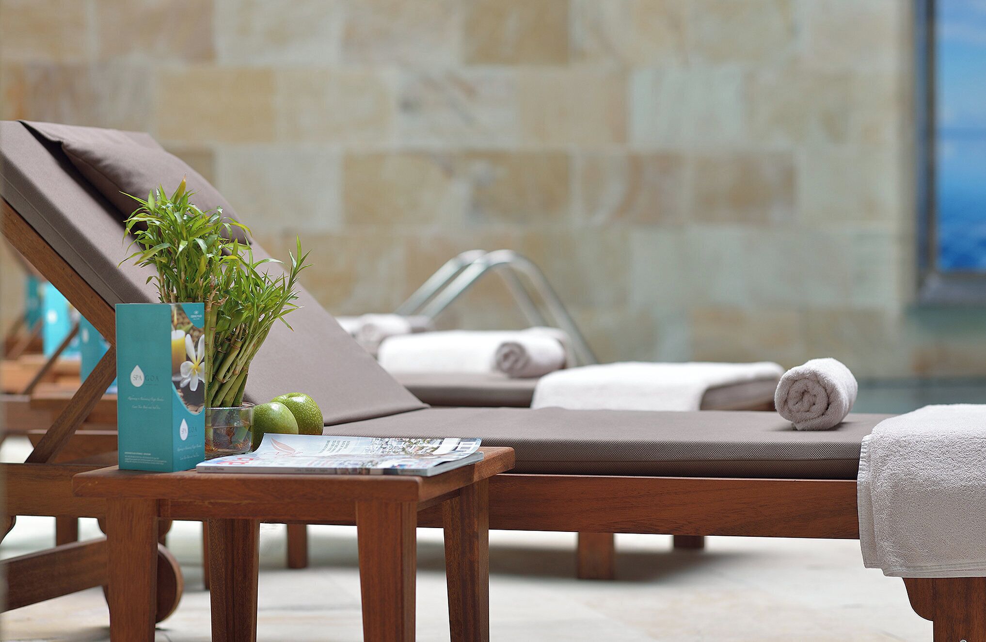 A spa area with wooden loungers, a small table with a plant and a magazine, and a tiled wall in the background.