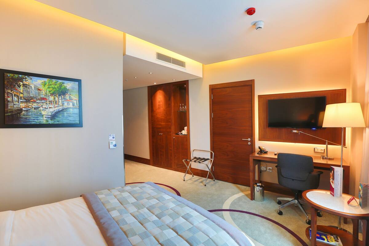 A hotel room with a bed, desk, chair, TV, and a painting of a canal scene.