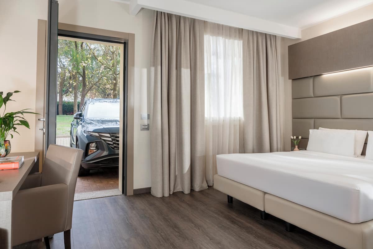 A hotel room with a bed, a desk, and a chair. There is a view of a car and trees outside the window.