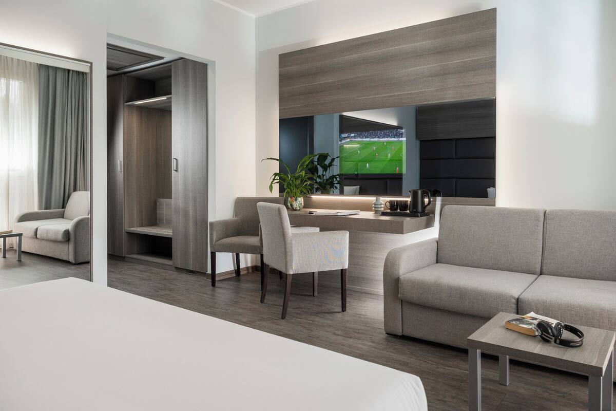 A modern hotel room with a large bed, a seating area with a sofa and chairs, a small table, a TV mounted on the wall, and a closet with a sliding door.