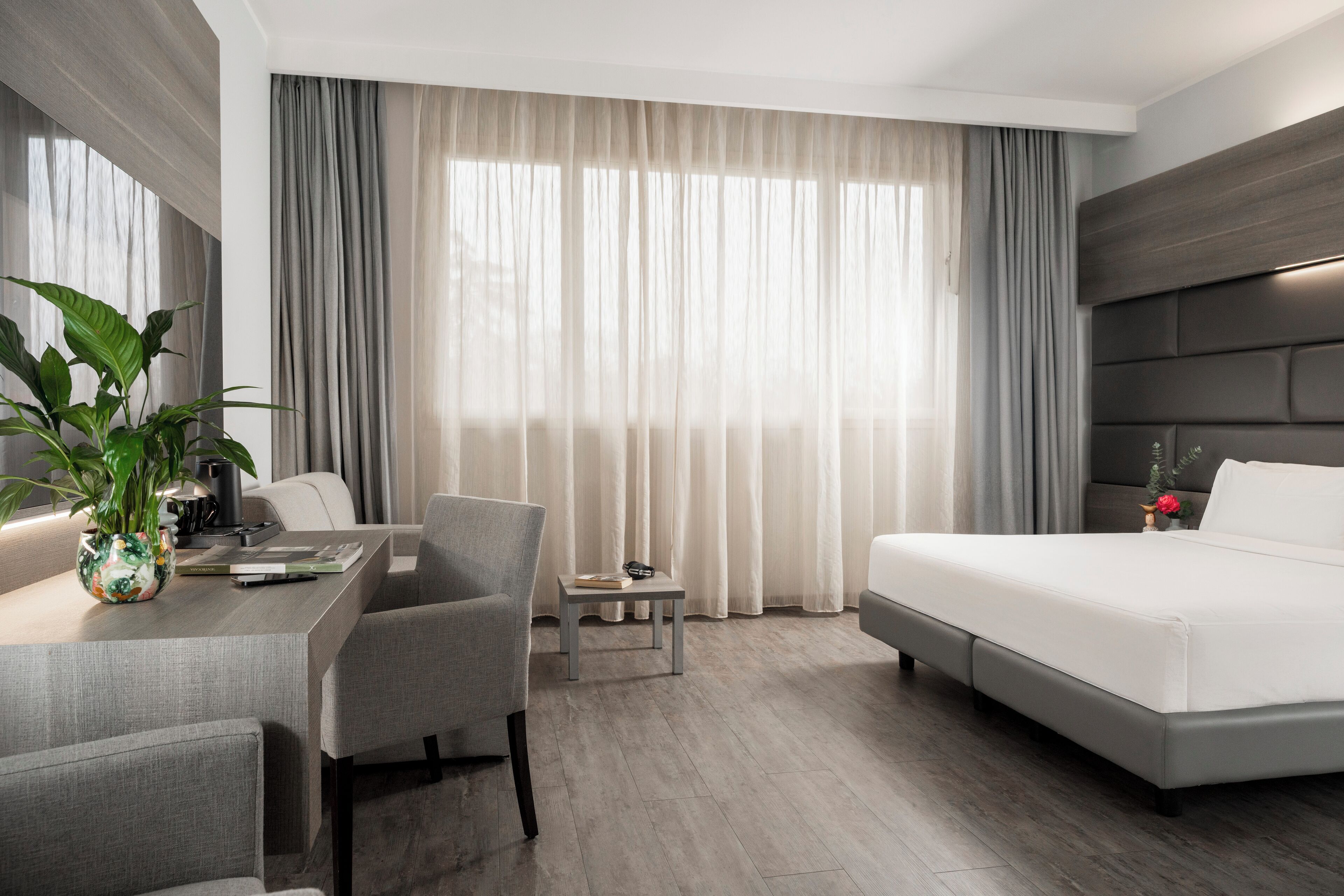 A modern hotel room with a bed, a desk, and a chair. There is a plant on the desk and a small table with a lamp.