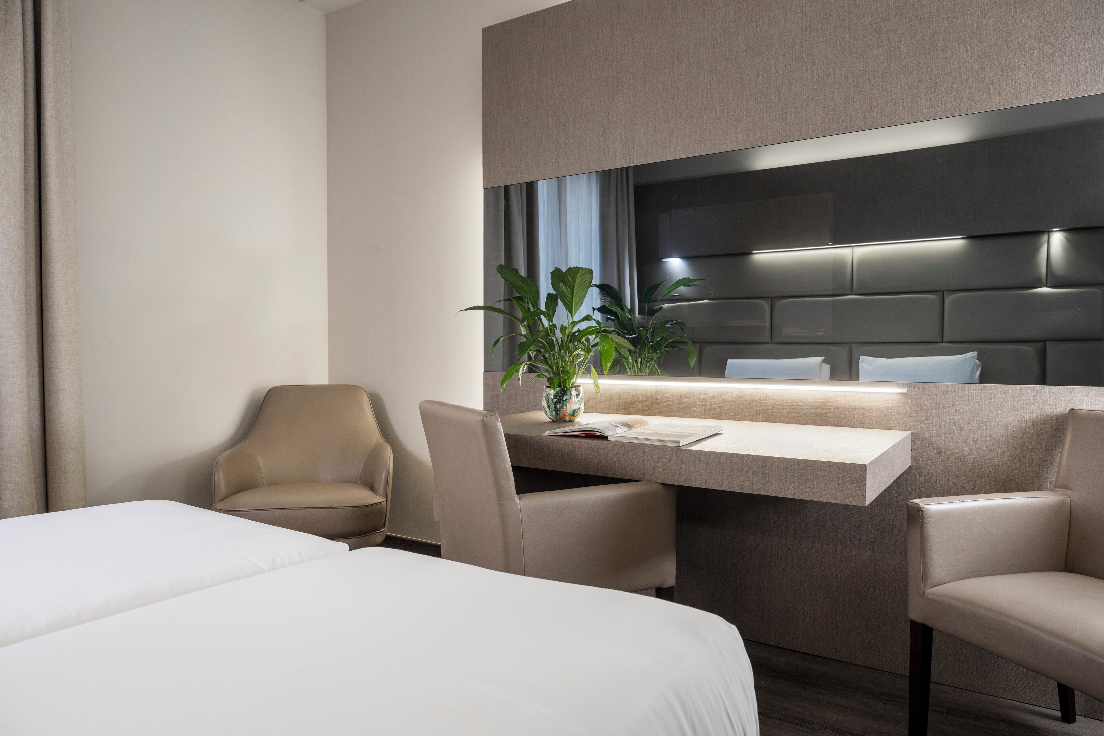 A modern hotel room with a bed, a desk, two chairs, a potted plant, and a large mirror.