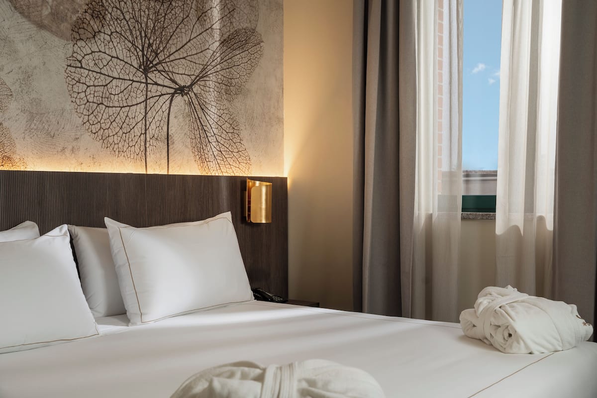 A hotel room with a bed, pillows, a headboard with a leaf design, a bedside lamp, a window with curtains, and a towel on the bed.