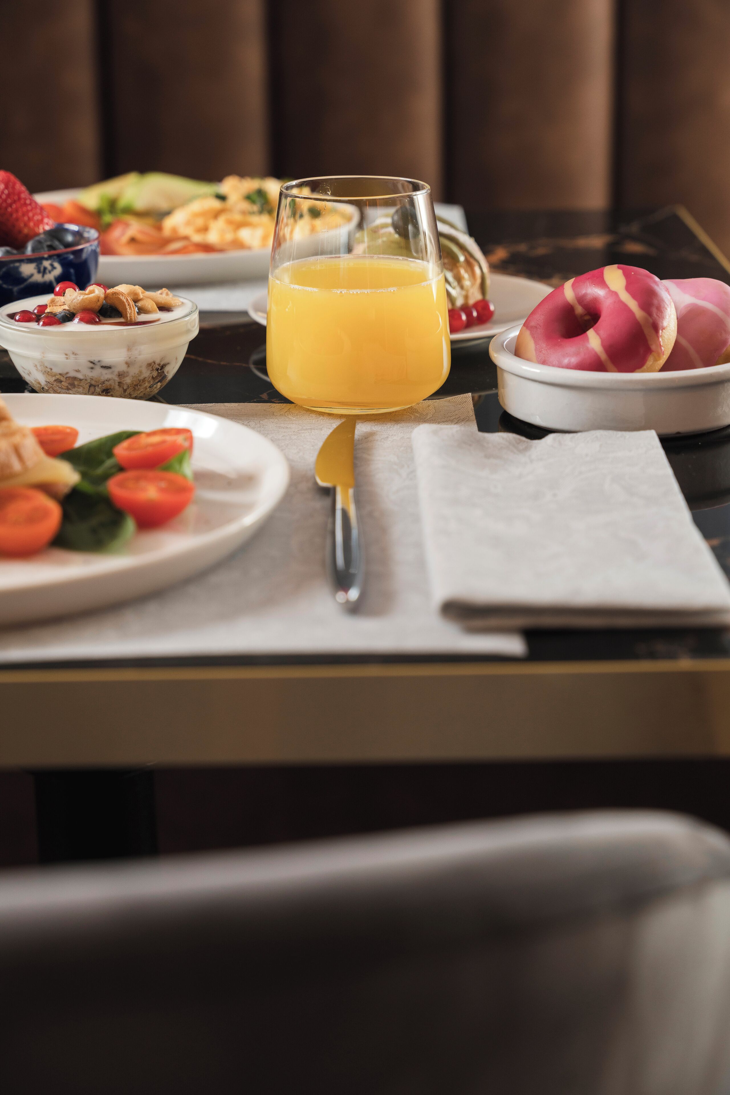 Una mesa de desayuno con un vaso de jugo de naranja, un plato de tomates en rodajas, un tazón de cereal y un plato de pasteles.