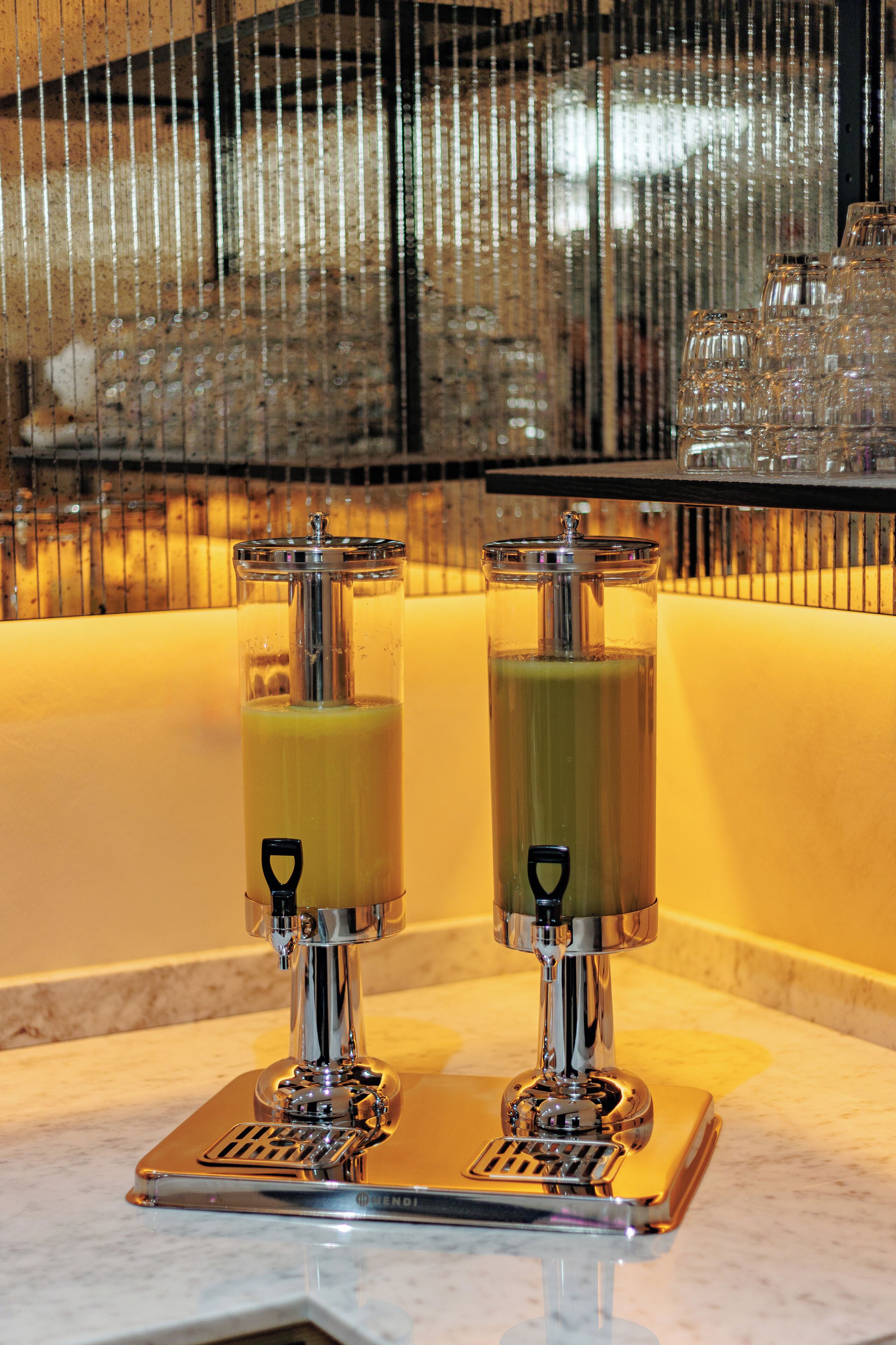 A bar area with two tall, clear dispensers filled with a green liquid, likely a cocktail or juice, on a reflective surface.