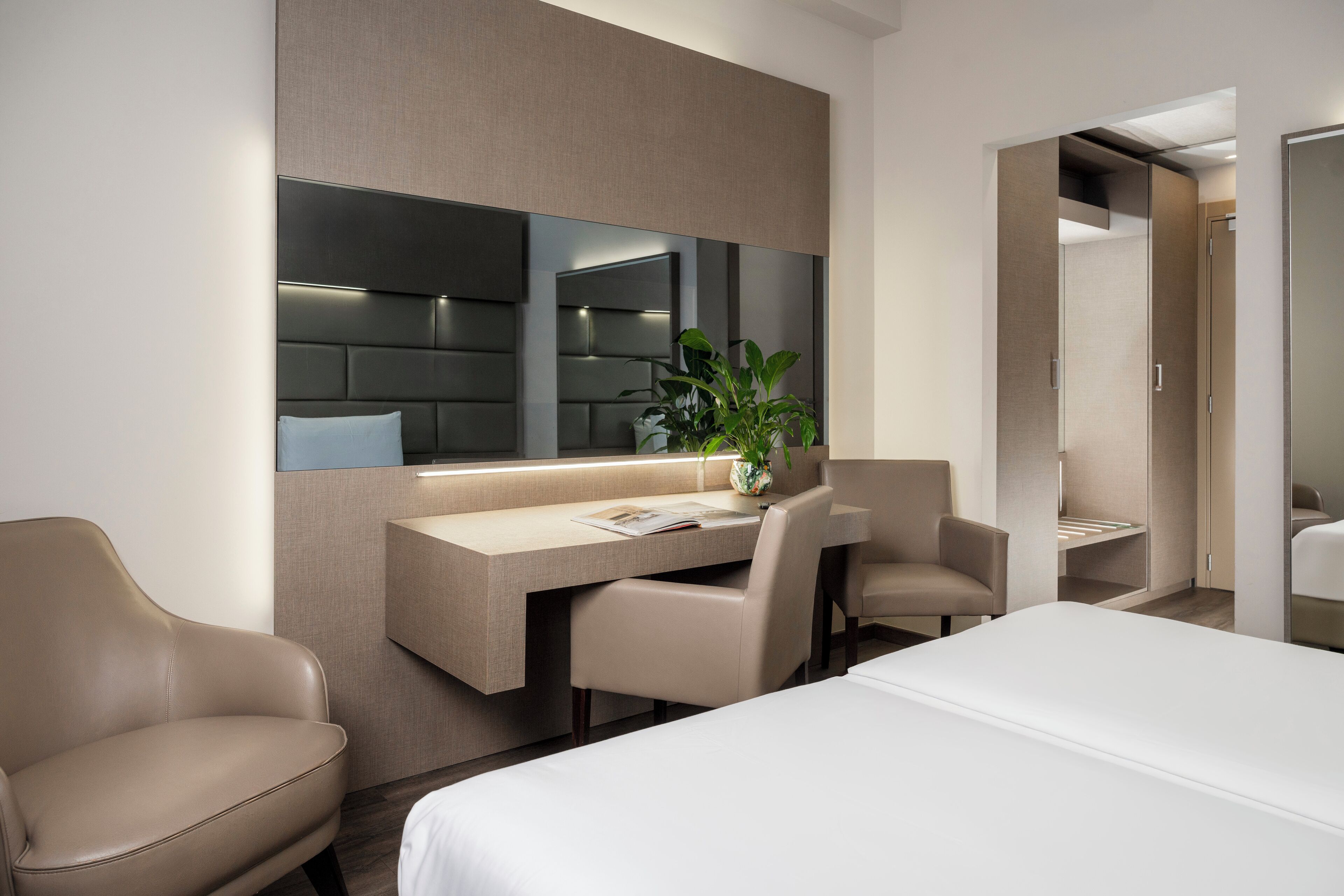A modern hotel room with a bed, a desk, two chairs, a large mirror, and a potted plant.