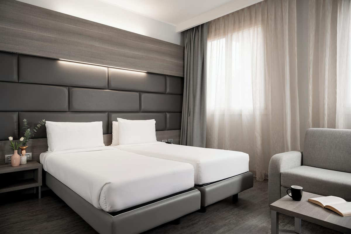 A modern hotel room with a large bed, a sofa, a bedside table, and a window with curtains.