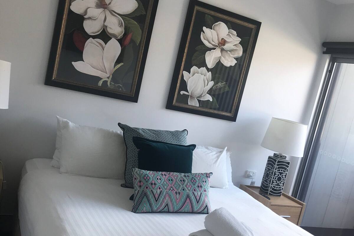 Una cama bien hecha con sábanas blancas, una toalla enrollada y cojines decorativos. Dos cuadros con motivos florales están colgados en la pared sobre la cama.