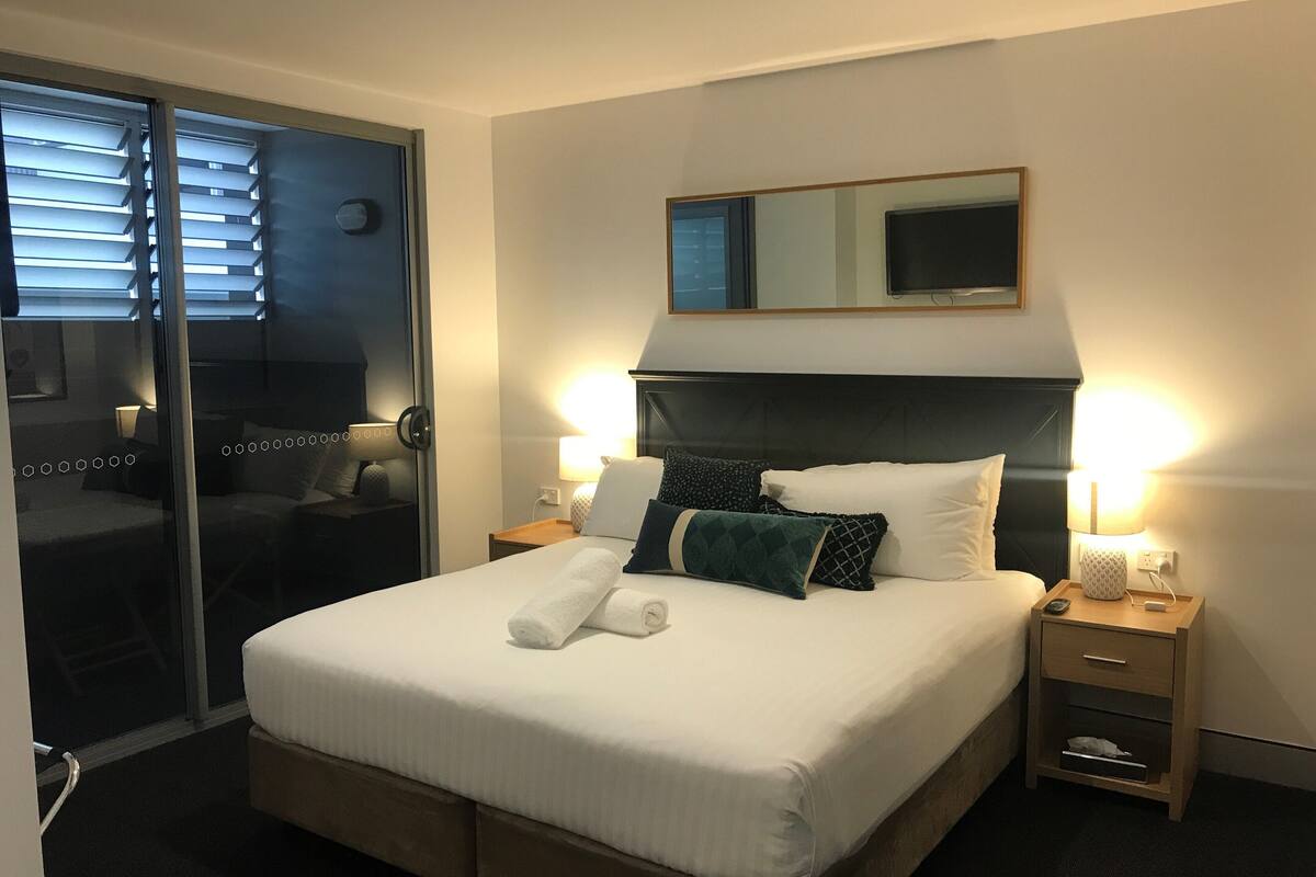 Una habitación de hotel con una cama grande, dos mesitas de noche con lámparas, un espejo sobre la cama y una puerta corredera que da a otra habitación.