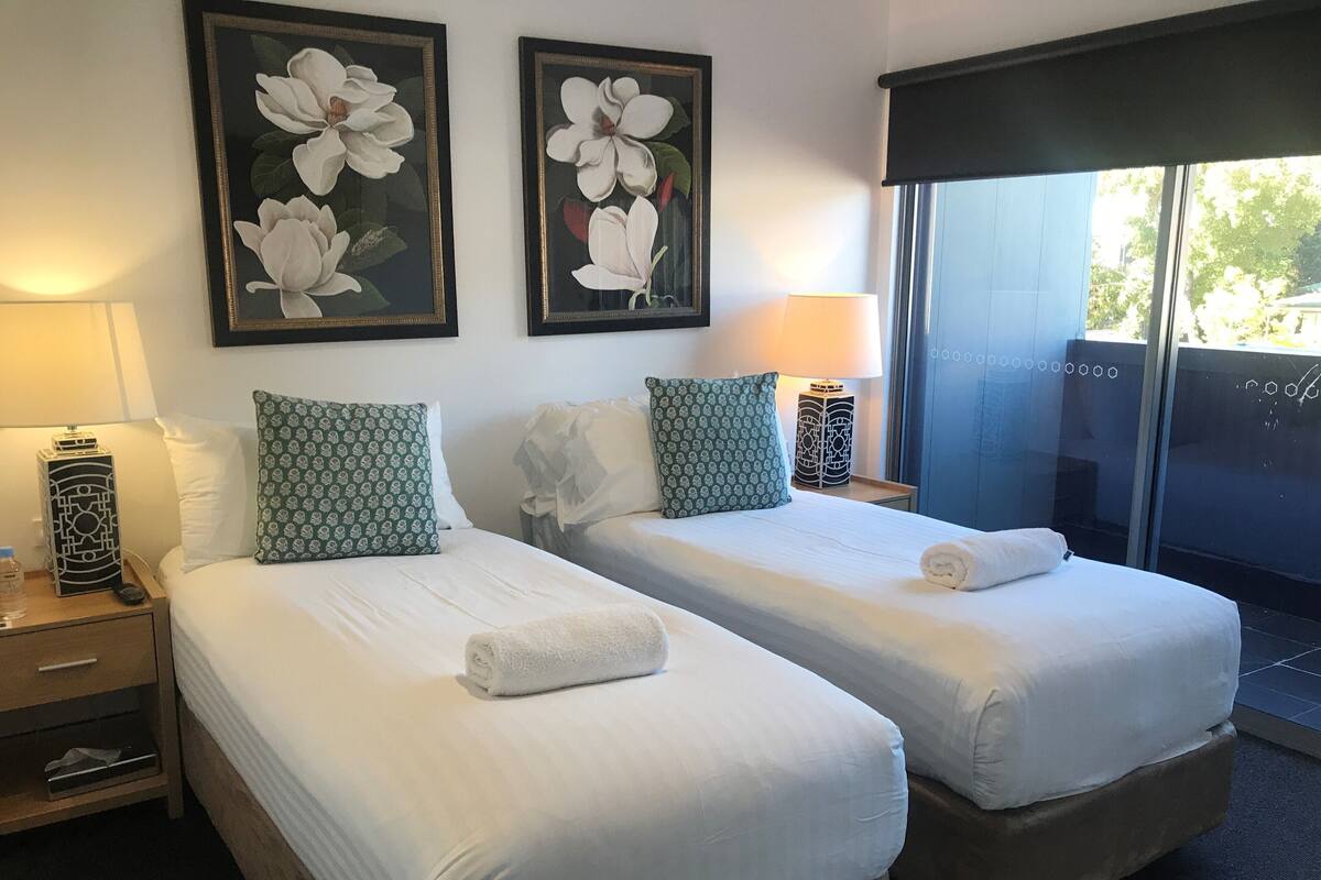 Habitación de hotel con dos camas, cada una con ropa de cama blanca y almohadas de diseño verde. En la pared hay cuadros con motivos florales, una mesita de noche con lámpara y un ventanal con vistas al exterior.