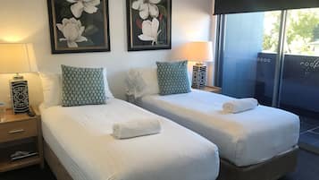 A hotel room with two beds, each with white linens and green patterned pillows. There are framed floral paintings on the wall, a bedside table with a lamp, and a large window with a view of the outdoors.