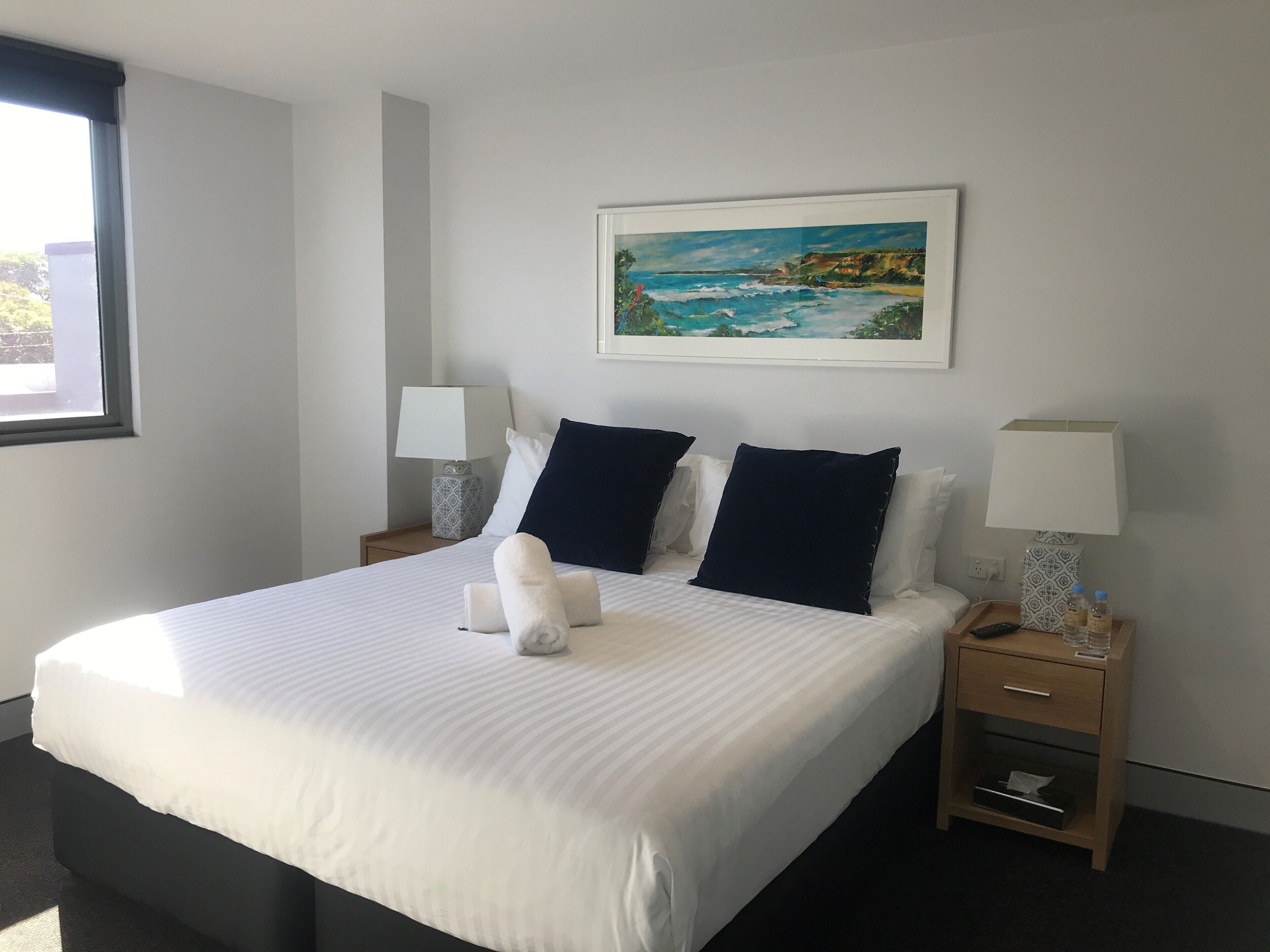 Una cama bien hecha con sábanas blancas y dos almohadas azul marino, un cuadro con una escena costera en la pared, y una mesita de noche con una lámpara y una botella.
