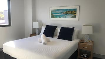 Una cama bien hecha con sábanas blancas y dos almohadas azul marino, un cuadro con una escena costera en la pared, y una mesita de noche con una lámpara y una botella.