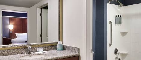 Une salle de bain d’hôtel avec un lavabo doté d’un plan de travail en granit, un grand miroir, une douche avec un pommeau de douche à main et des toilettes.