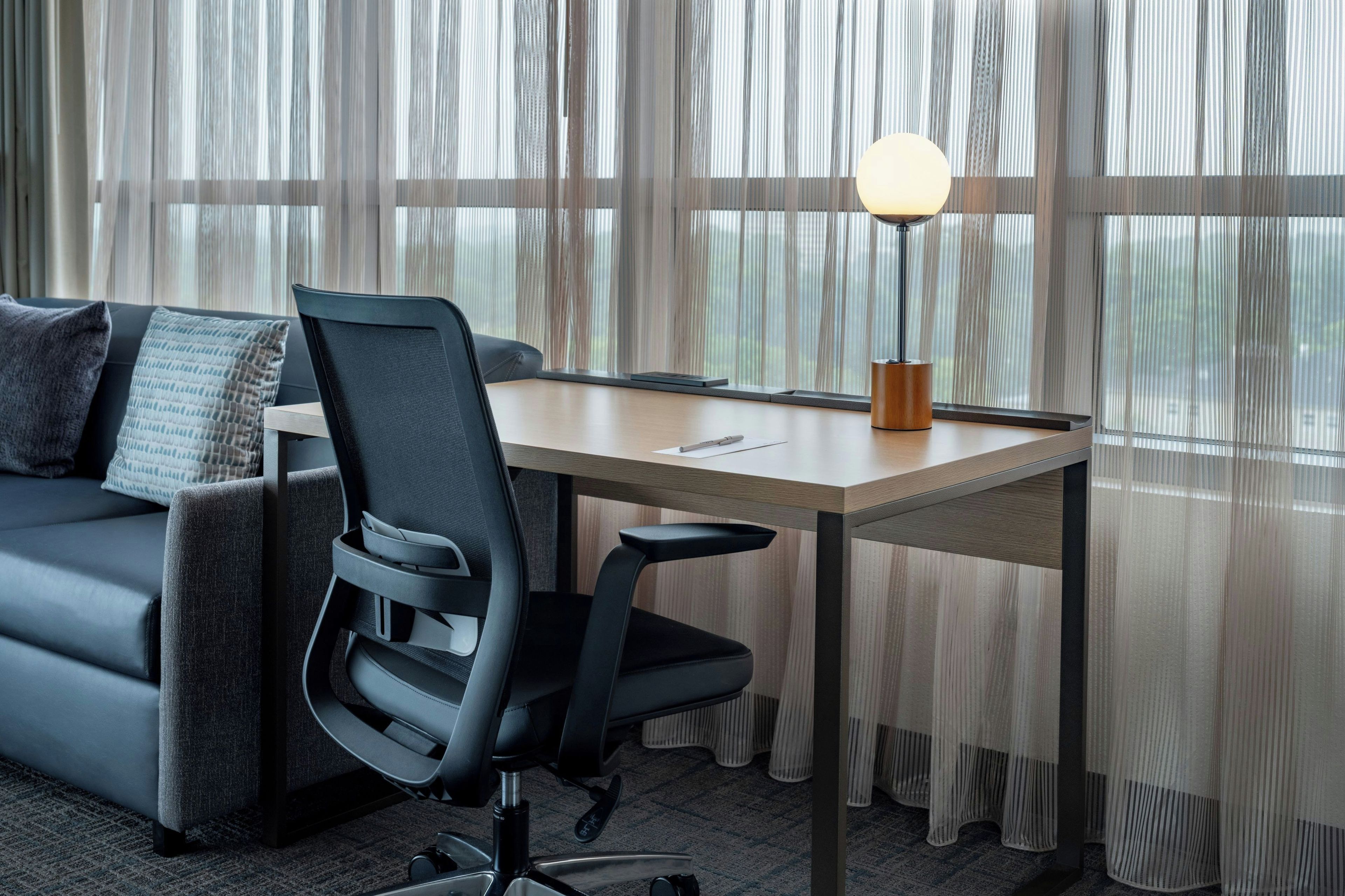 Ein moderner Arbeitsbereich mit grauem Sofa, schwarzem Bürostuhl, Holztisch und Lampe.