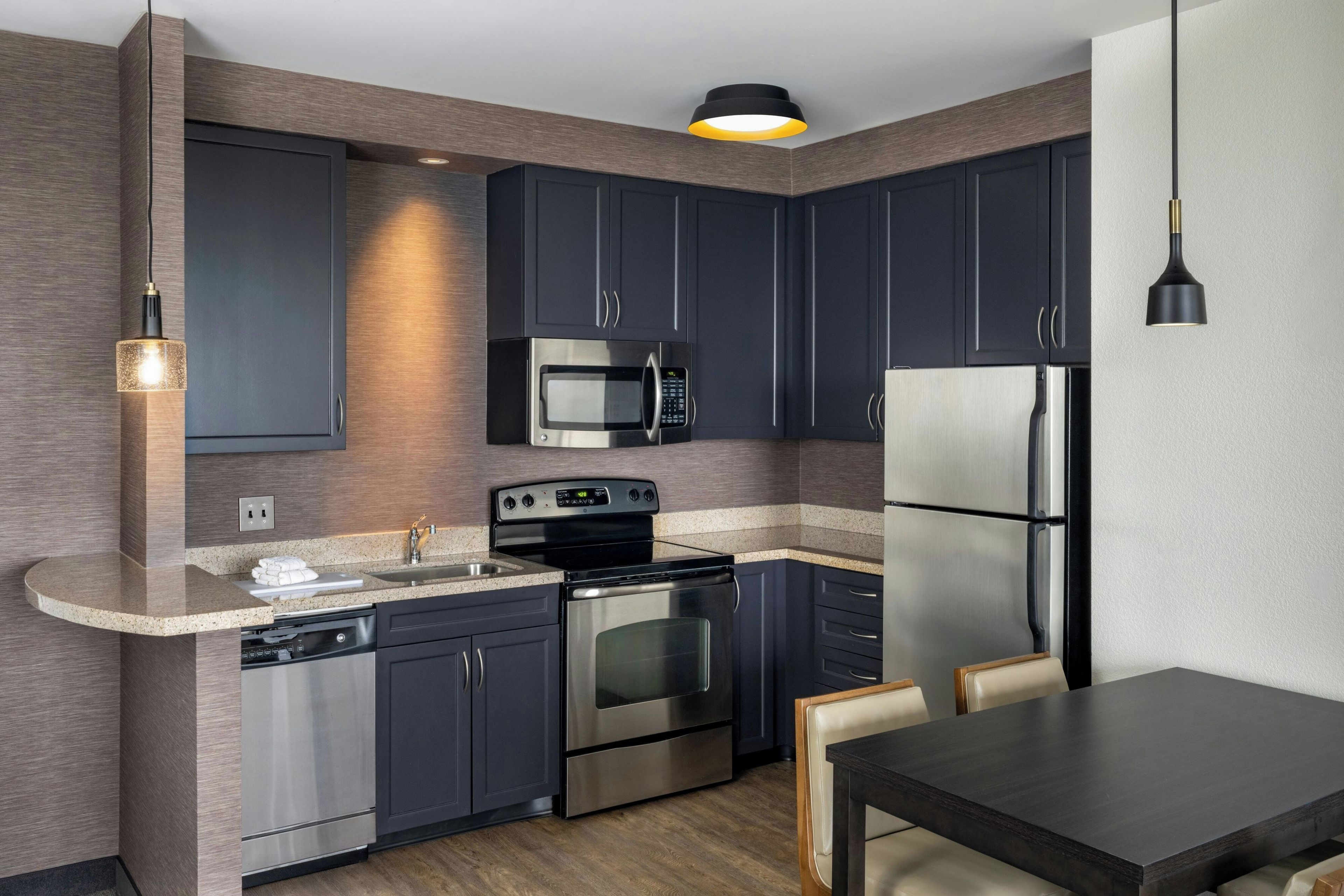 Una cocina moderna con armarios oscuros, electrodomésticos de acero inoxidable, horno empotrado, microondas y refrigerador.