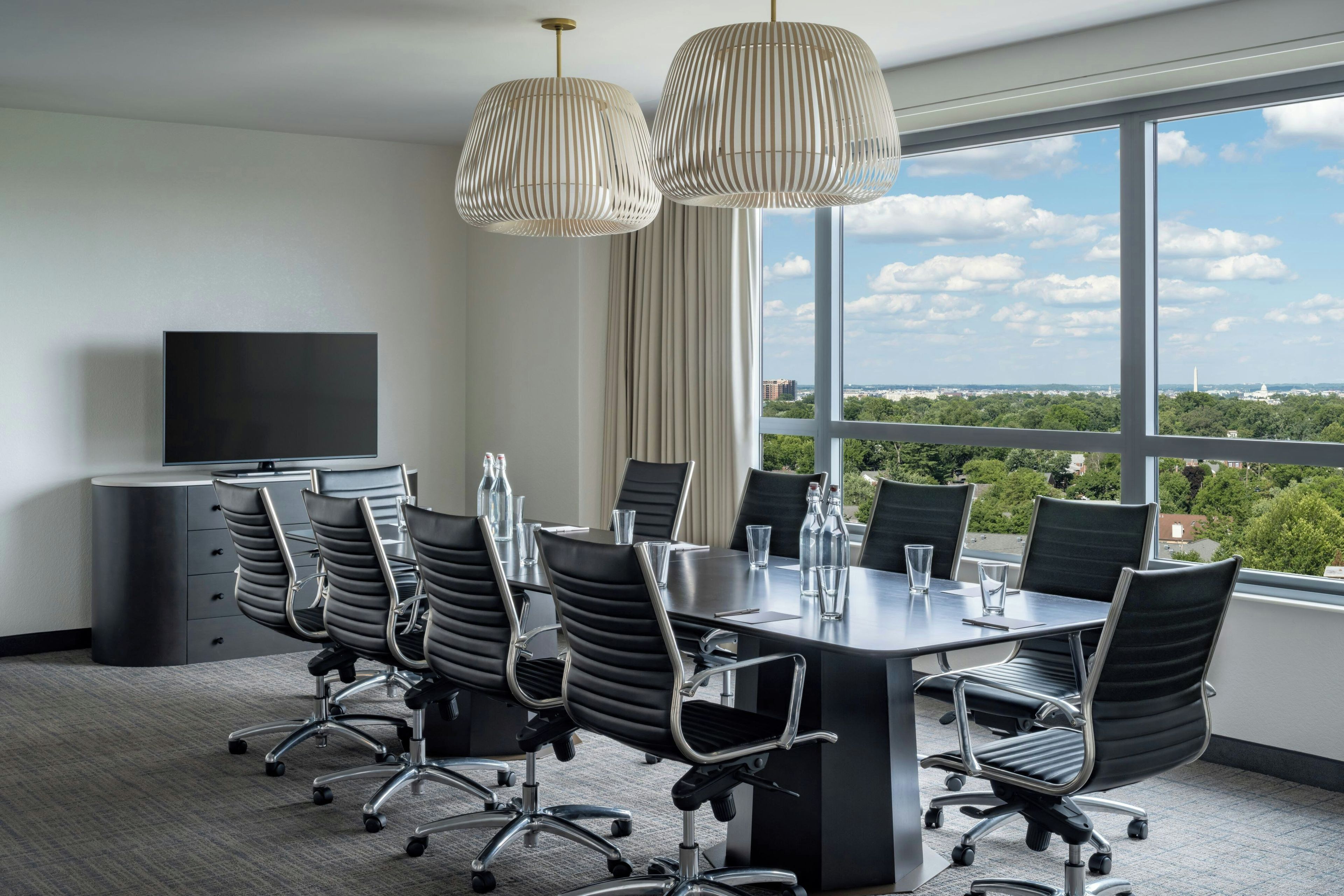 Un salón de conferencias con una gran mesa, sillas, un televisor y vistas a un paisaje urbano.