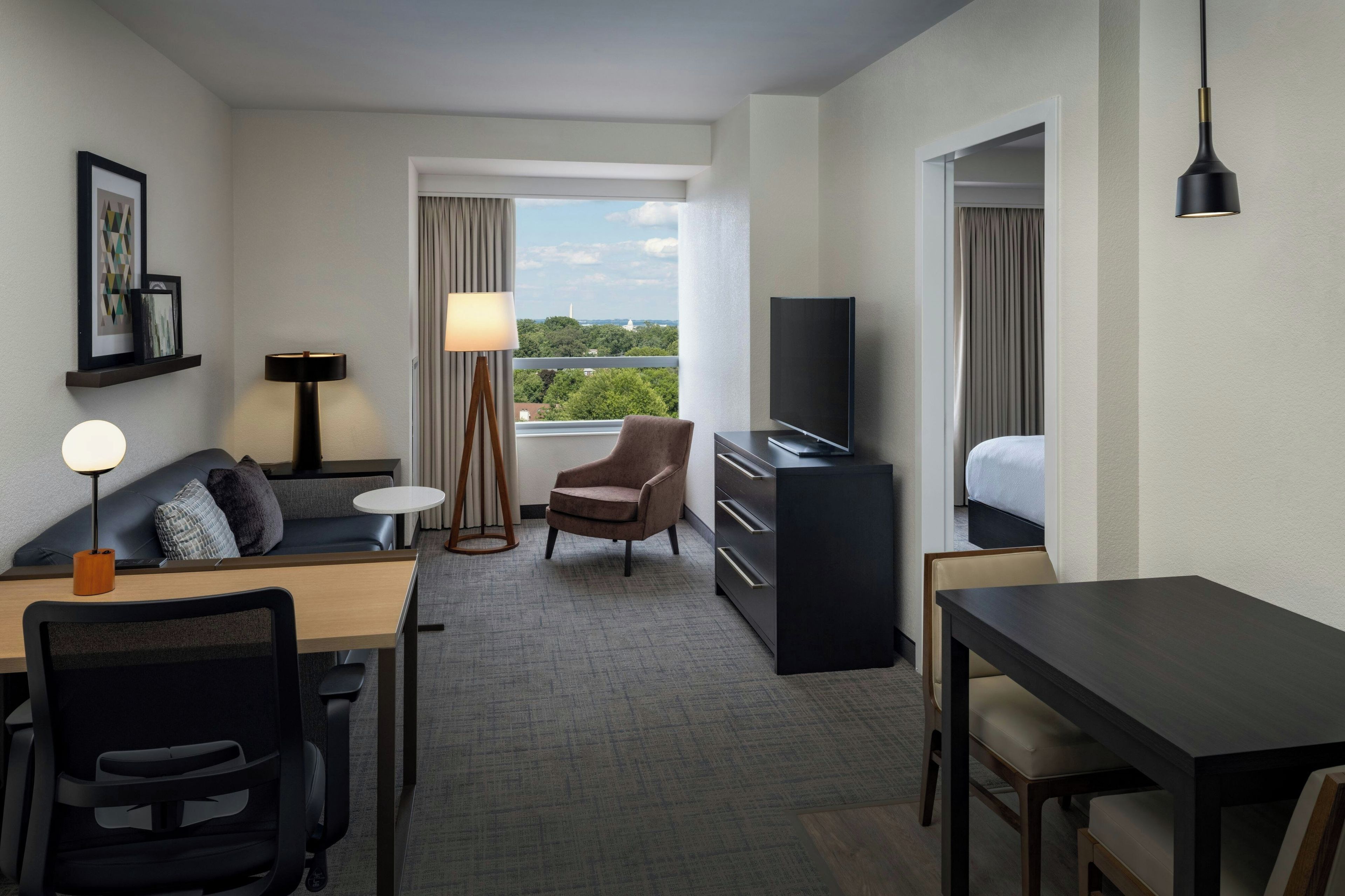 A hotel room with a desk, chair, sofa, TV, and a view of the cityscape.