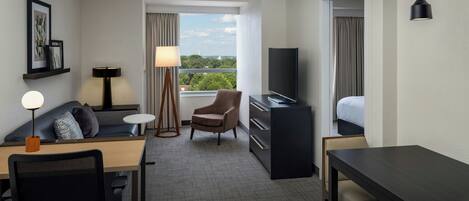 A hotel room with a desk, chair, sofa, TV, and a view of the cityscape.