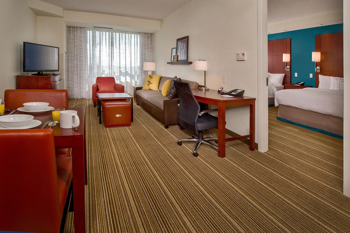 Ein Hotelzimmer mit Bett, Schreibtisch, Stuhl, Fernseher und einem Essbereich mit Tisch und Stühlen.