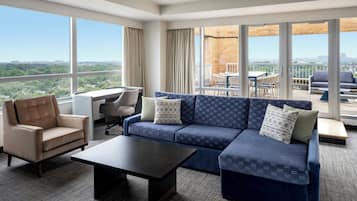 A modern hotel room with a large window, a sofa, a chair, a desk, and a balcony with outdoor furniture.