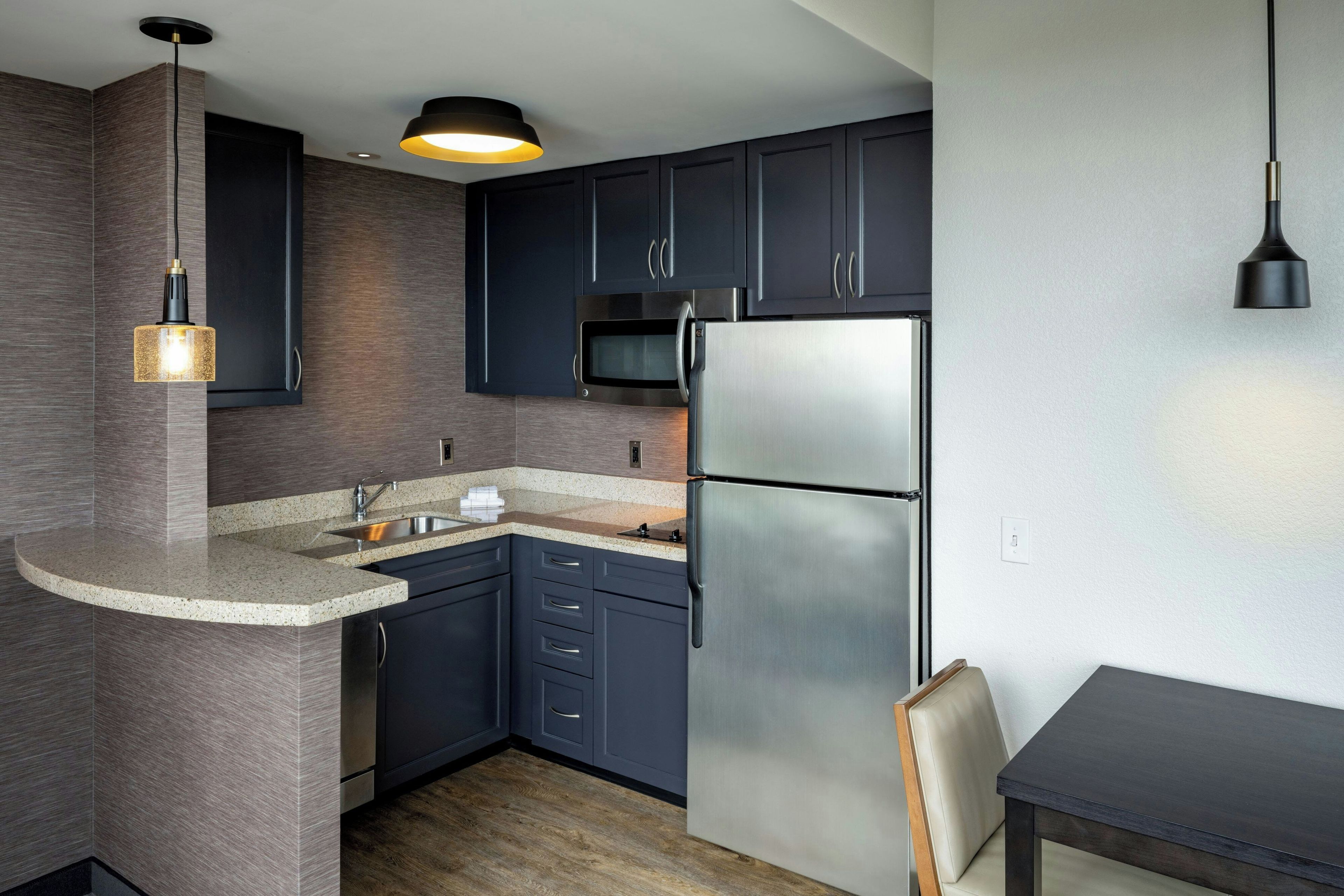 Una cocina moderna con armarios oscuros, un refrigerador de acero inoxidable, una encimera de granito y una lámpara colgante.