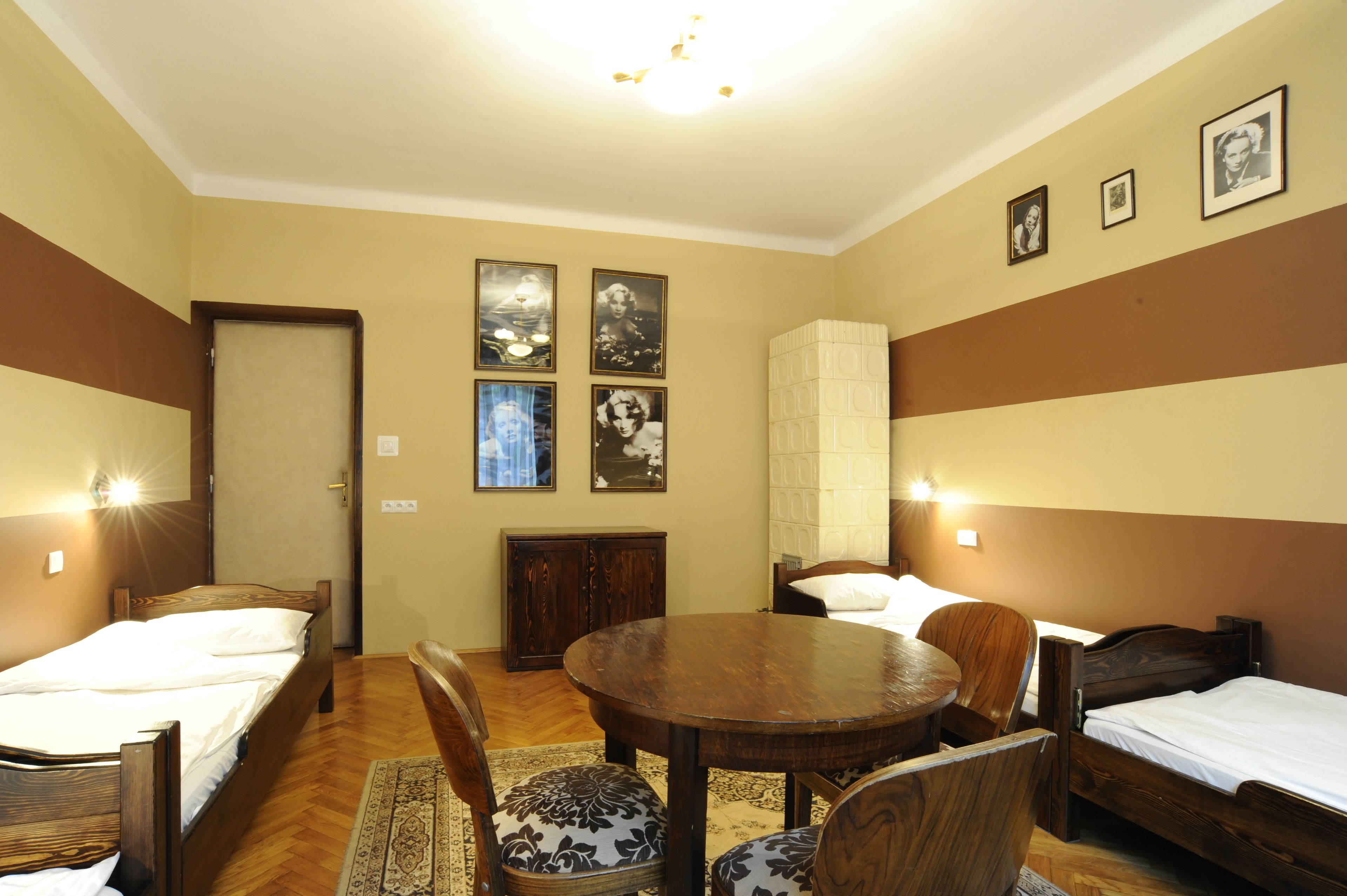 A room with two single beds, a wooden dining table, and a cabinet. There are framed pictures on the wall and a door with a light above it.