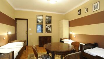 A room with two single beds, a wooden dining table, and a cabinet. There are framed pictures on the wall and a door with a light above it.