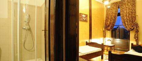 A room with a glass-enclosed shower, a bed with a wooden headboard, a small table, and a window with curtains.