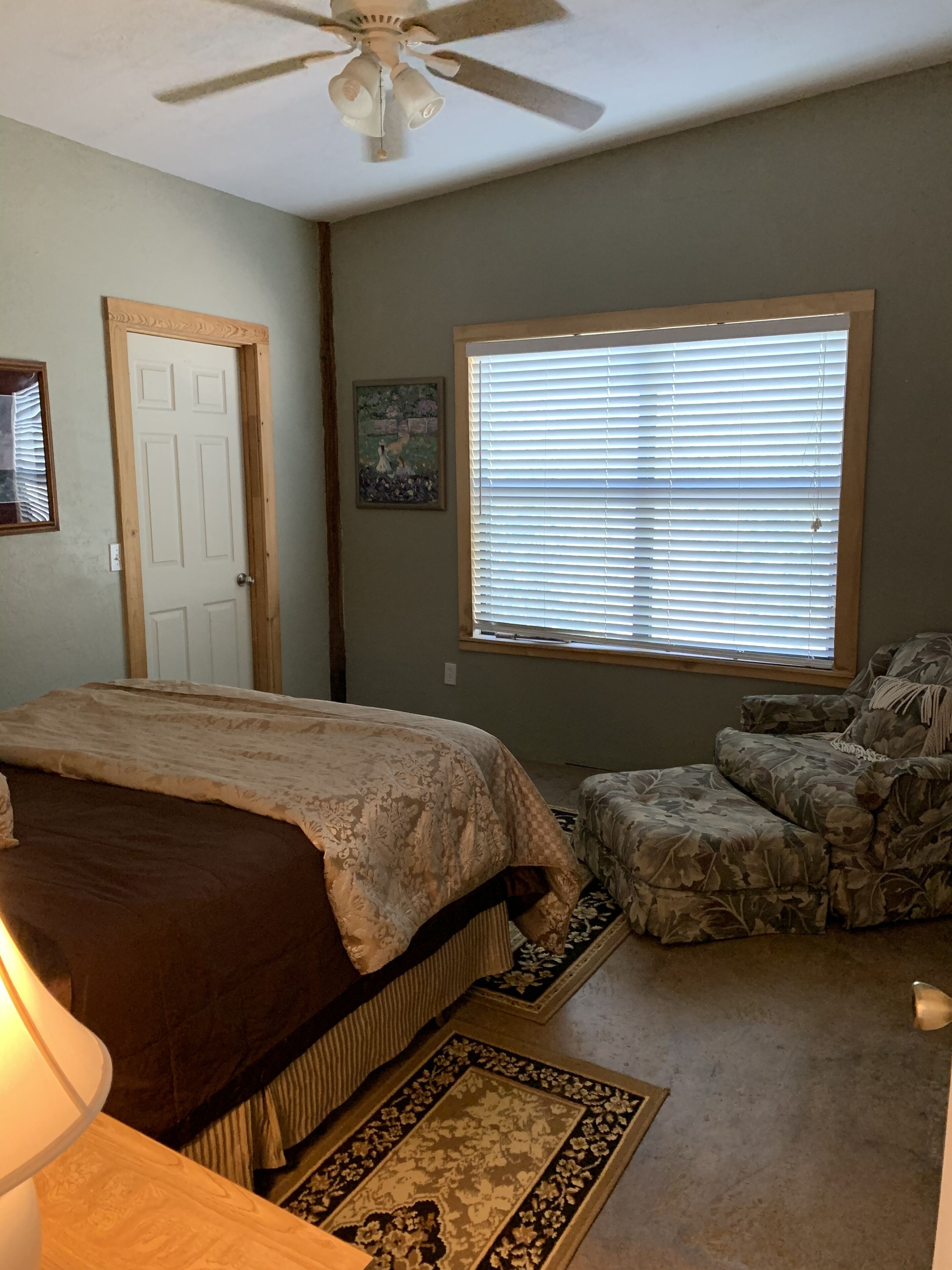 Une chambre à coucher comprenant un lit, une chaise, une fenêtre avec des stores, un ventilateur de plafond et une porte.