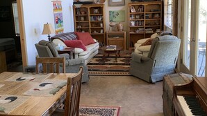 Amplia sala de estar con techo de madera, una mesa de comedor con sillas, un sofá, un piano y dos grandes estanterías llenas de libros y objetos decorativos.