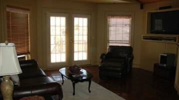 A living room with a sofa, armchair, and coffee table. There are two windows with blinds, a television, and a fireplace.