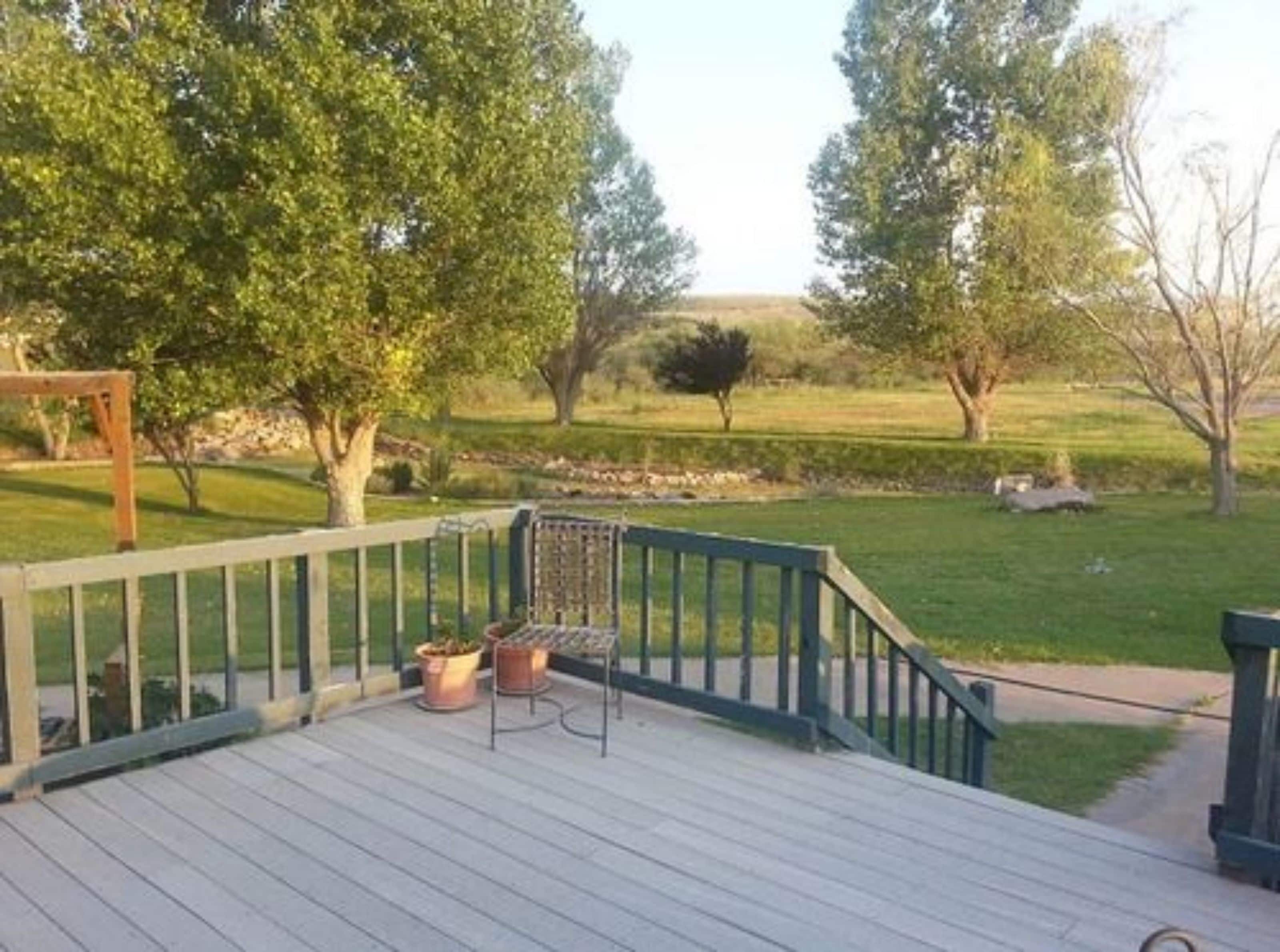 A wooden deck with a metal railing, potted plants, and a view of a grassy landscape with trees.