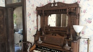Un piano droit ancien, avec un miroir au-dessus, une lampe et un papier peint à motifs floraux.