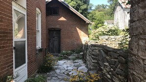 Un étroit sentier de pierres mène à une maison en briques, dotée d’une porte en bois sombre et d’une fenêtre à cadre blanc.