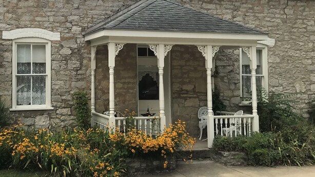 Un bâtiment en pierre doté d’un porche blanc et d’un petit jardin agrémenté de fleurs jaunes.
