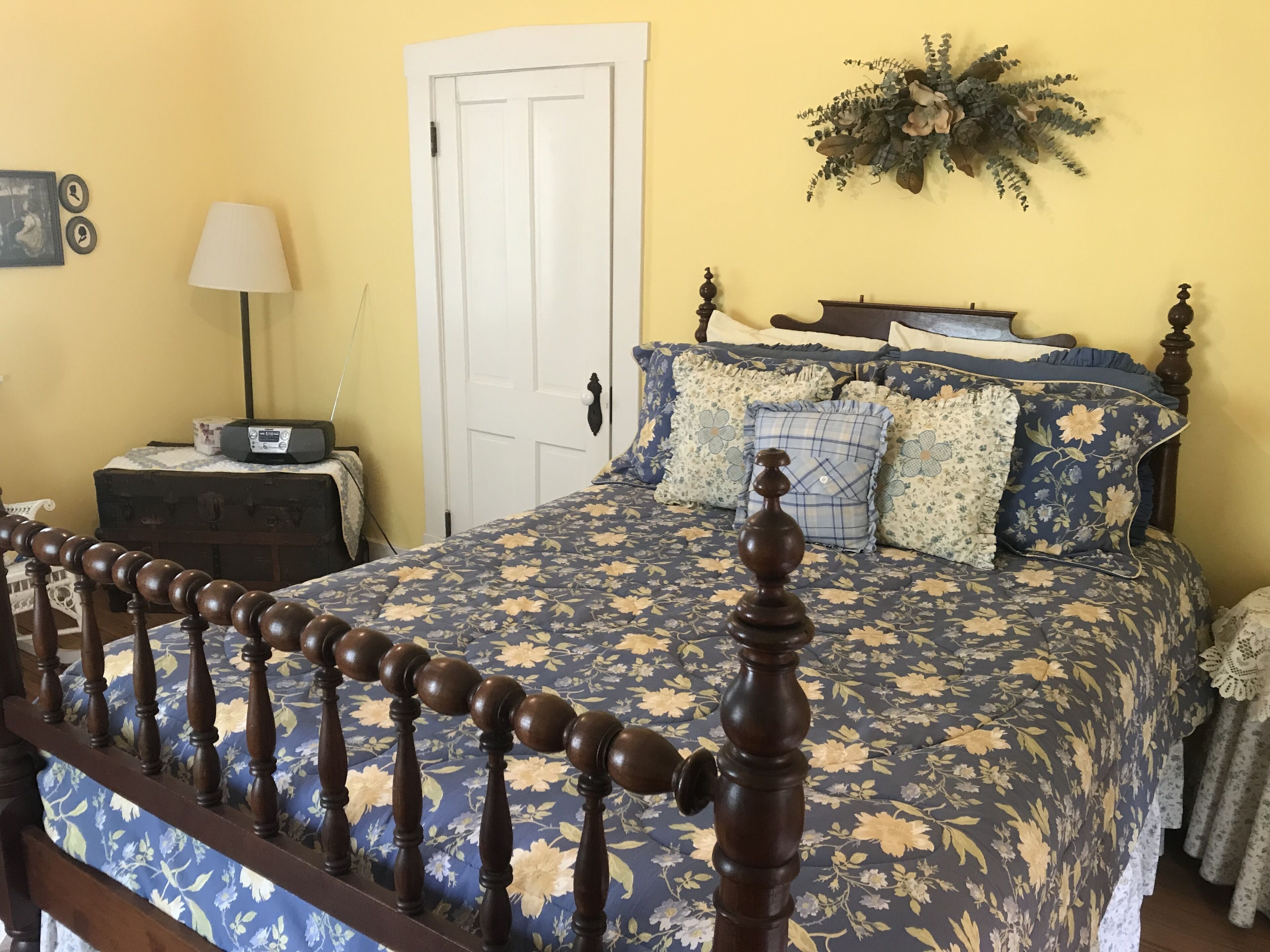 A bedroom with a floral-patterned bedspread, a wooden headboard, a bedside table with a lamp, and a door with a decorative wreath.