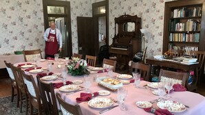 Une salle à manger avec une longue table dressée pour un repas, entourée de chaises recouvertes de housses en dentelle. On aperçoit un piano et une bibliothèque au fond de la pièce.