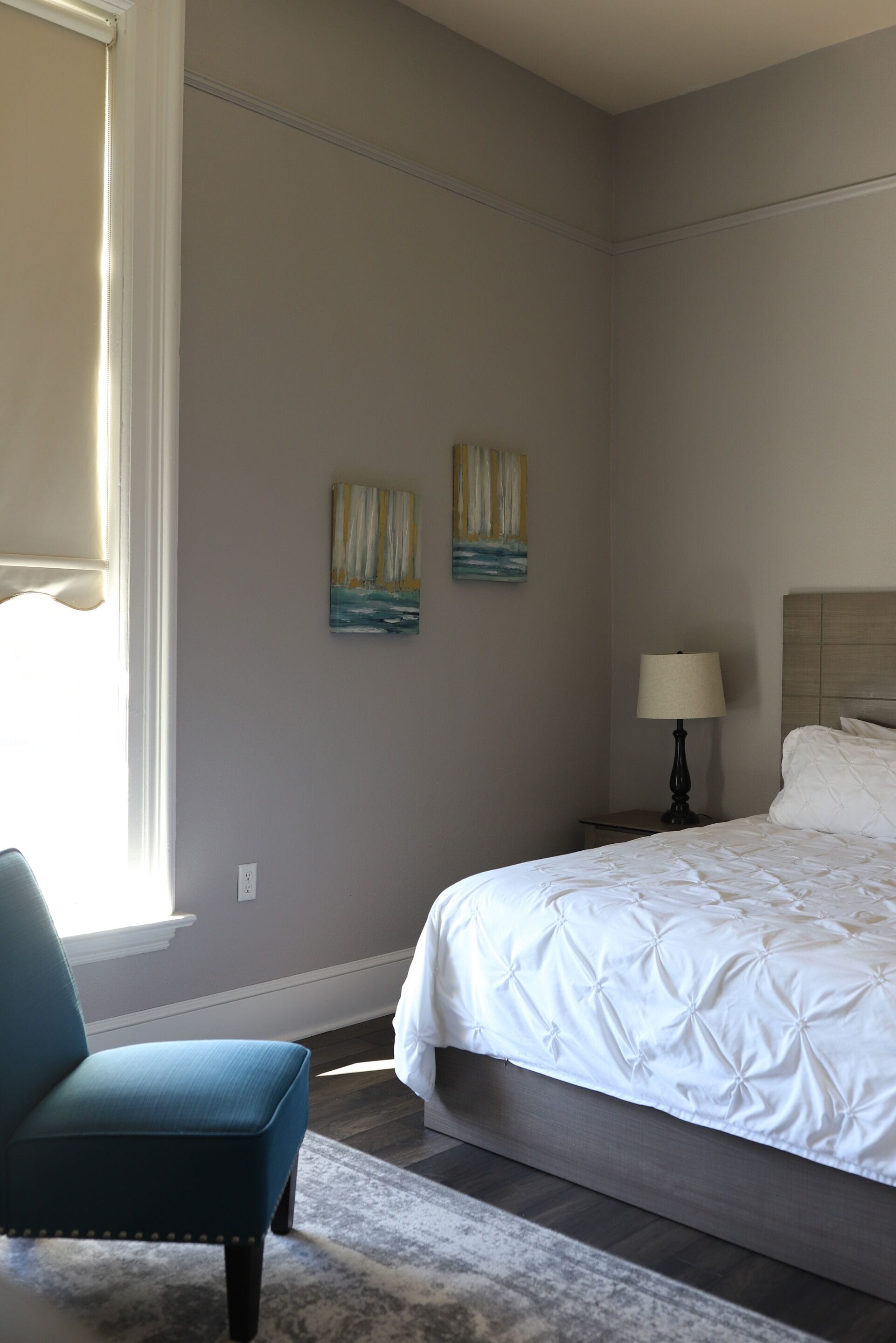 A bedroom with a bed, a chair, two paintings, a nightstand with a lamp, and a window with blinds.