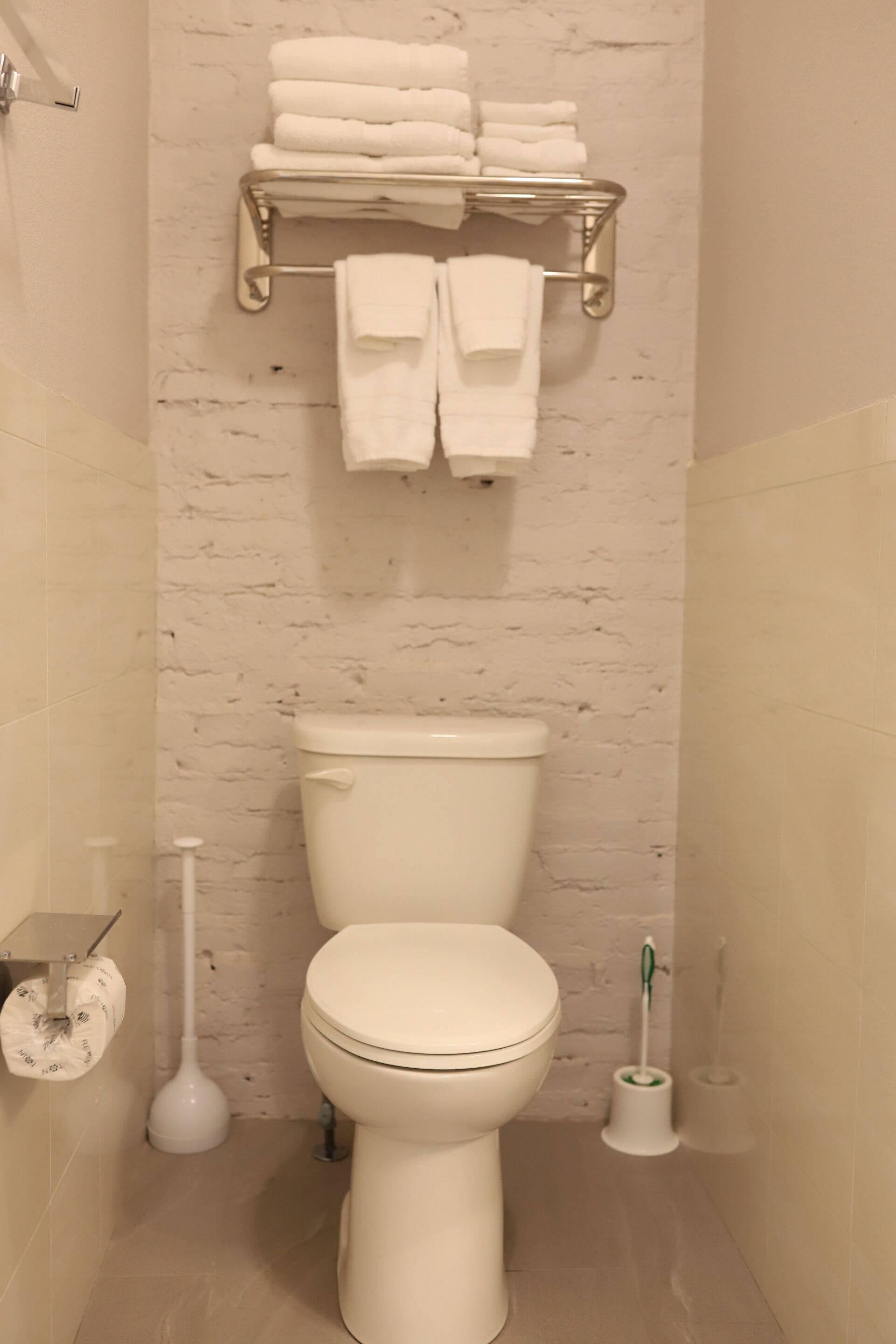 A bathroom with a white toilet, a towel rack with folded towels, a toilet brush, and a toilet paper holder.