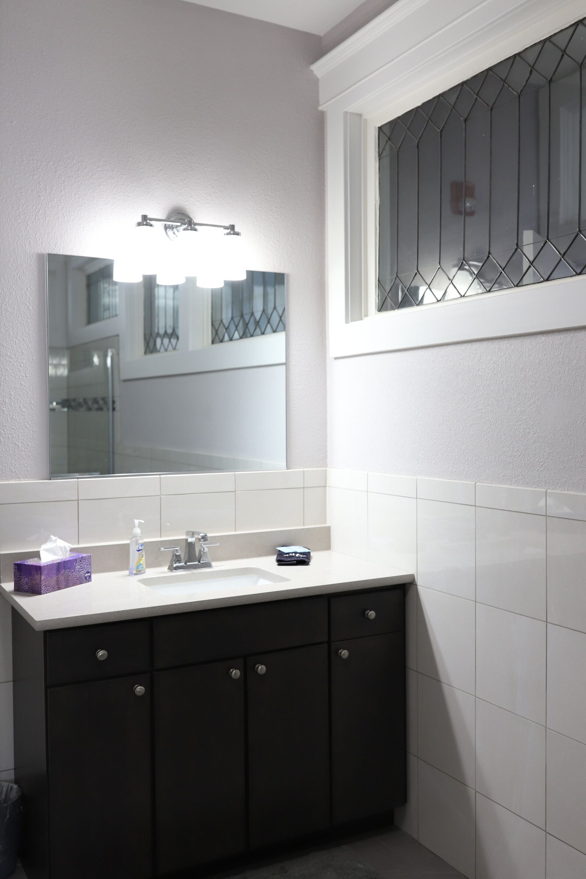 Ein Badezimmer mit einem weißen Waschbecken, dunklen Schränken, einem Spiegel und einem Fenster mit einem Metallgitter.