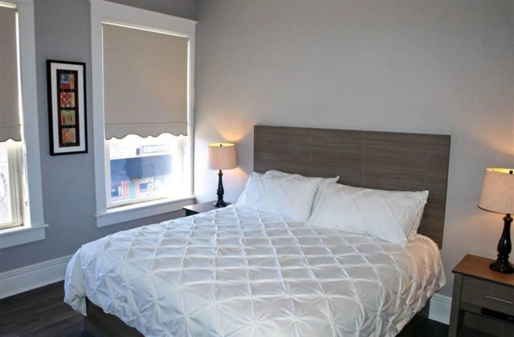 Ein ordentlich bezogenes Bett mit weißer Decke, zwei Nachttischlampen und ein Fenster mit Jalousien.