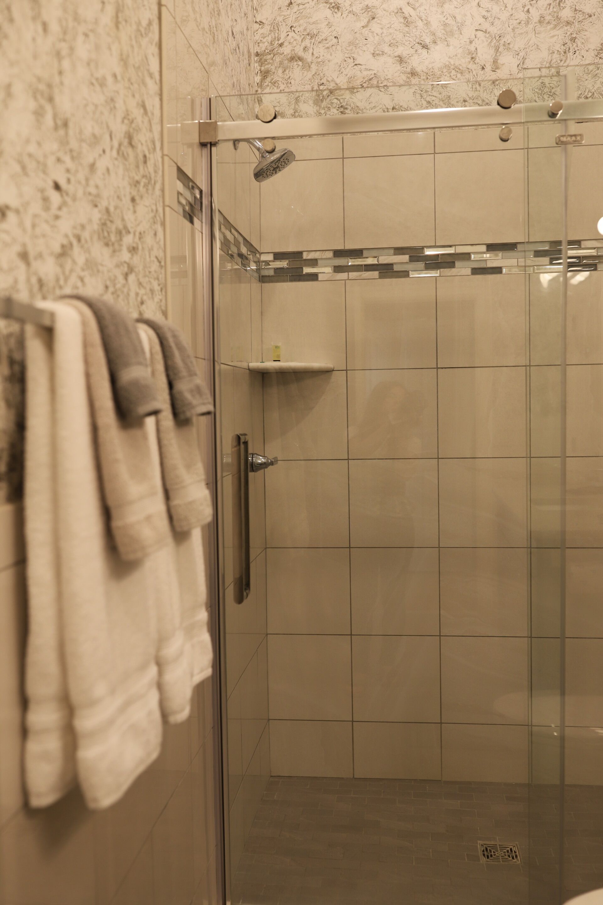 A glass-enclosed shower with a tiled wall and a towel rack with multiple towels.