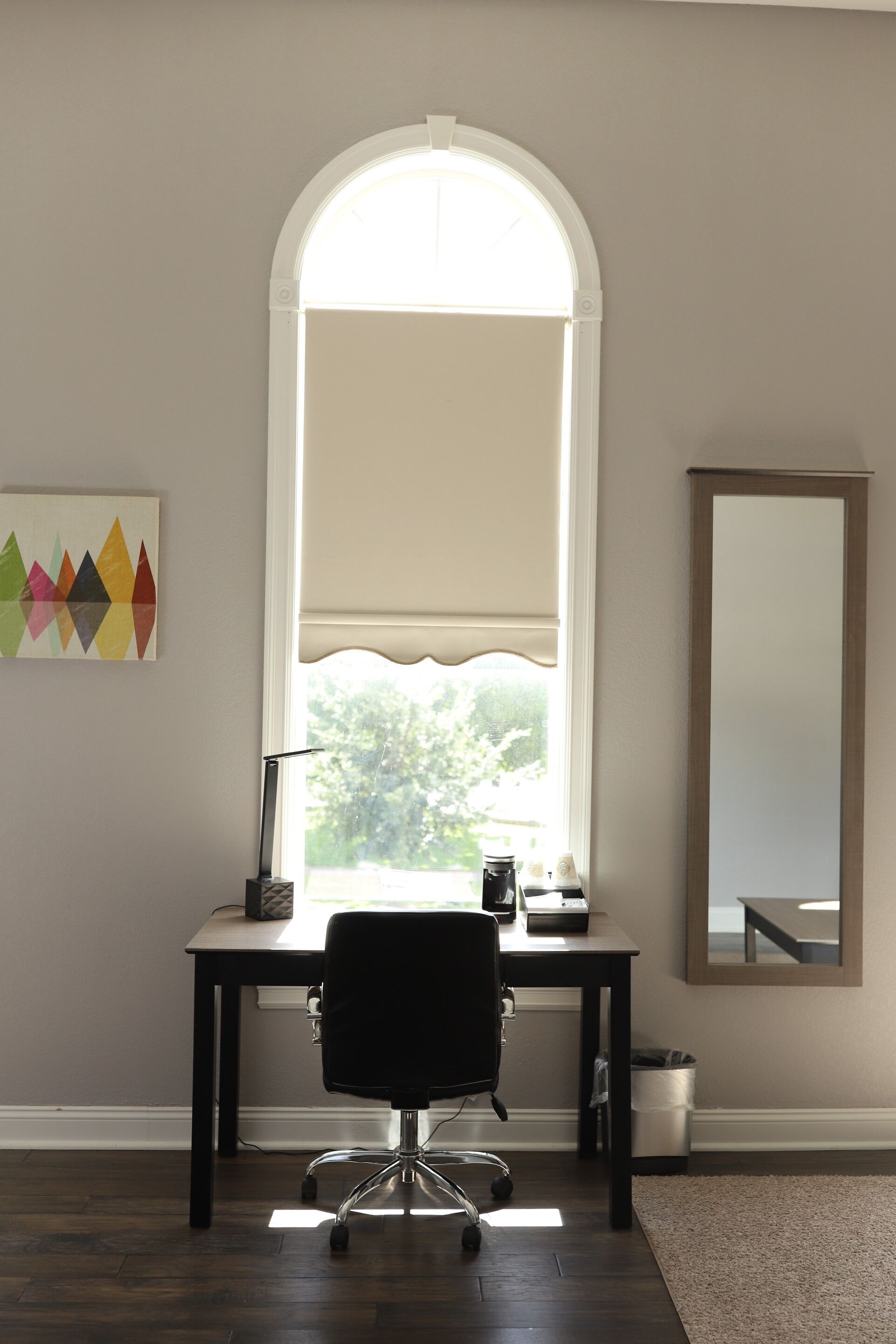 Una habitación con escritorio, silla, ventana con persianas, espejo y una obra de arte colorida en la pared.