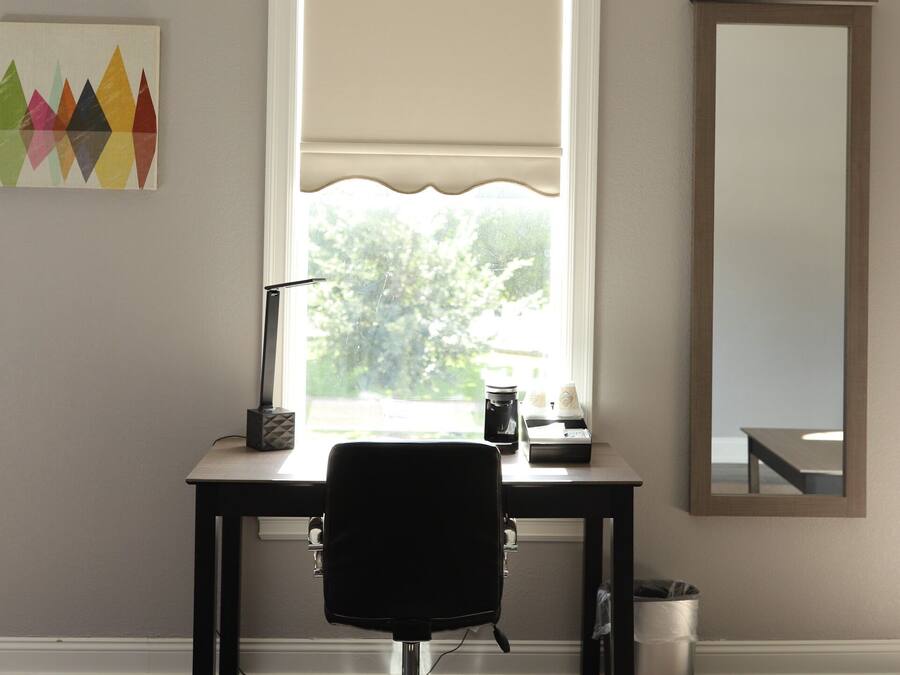 Una habitación con escritorio, silla, ventana con persianas, espejo y una obra de arte colorida en la pared.
