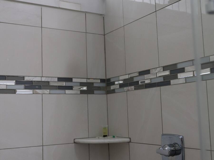 A bathroom with white tiles, a shower with a glass door, a small shelf, and a window with bars.