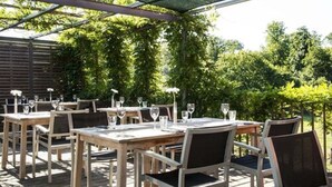 An outdoor dining area with wooden tables and chairs, covered by a green canopy, surrounded by lush greenery. - Hotel Simon's Plaza (Grevenmacher)