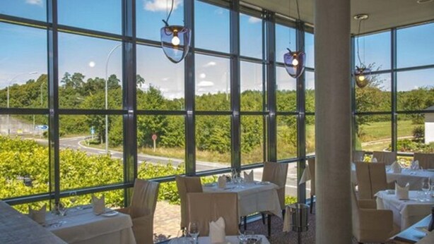 A restaurant with large windows offering a view of greenery and a road. Tables are set with white tablecloths and candles. - Hotel Simon's Plaza (Grevenmacher)