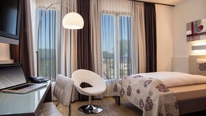 A hotel room with a bed, a desk with a laptop, a chair, a large window with curtains, and a floor lamp. - Hotel Simon's Plaza (Grevenmacher)