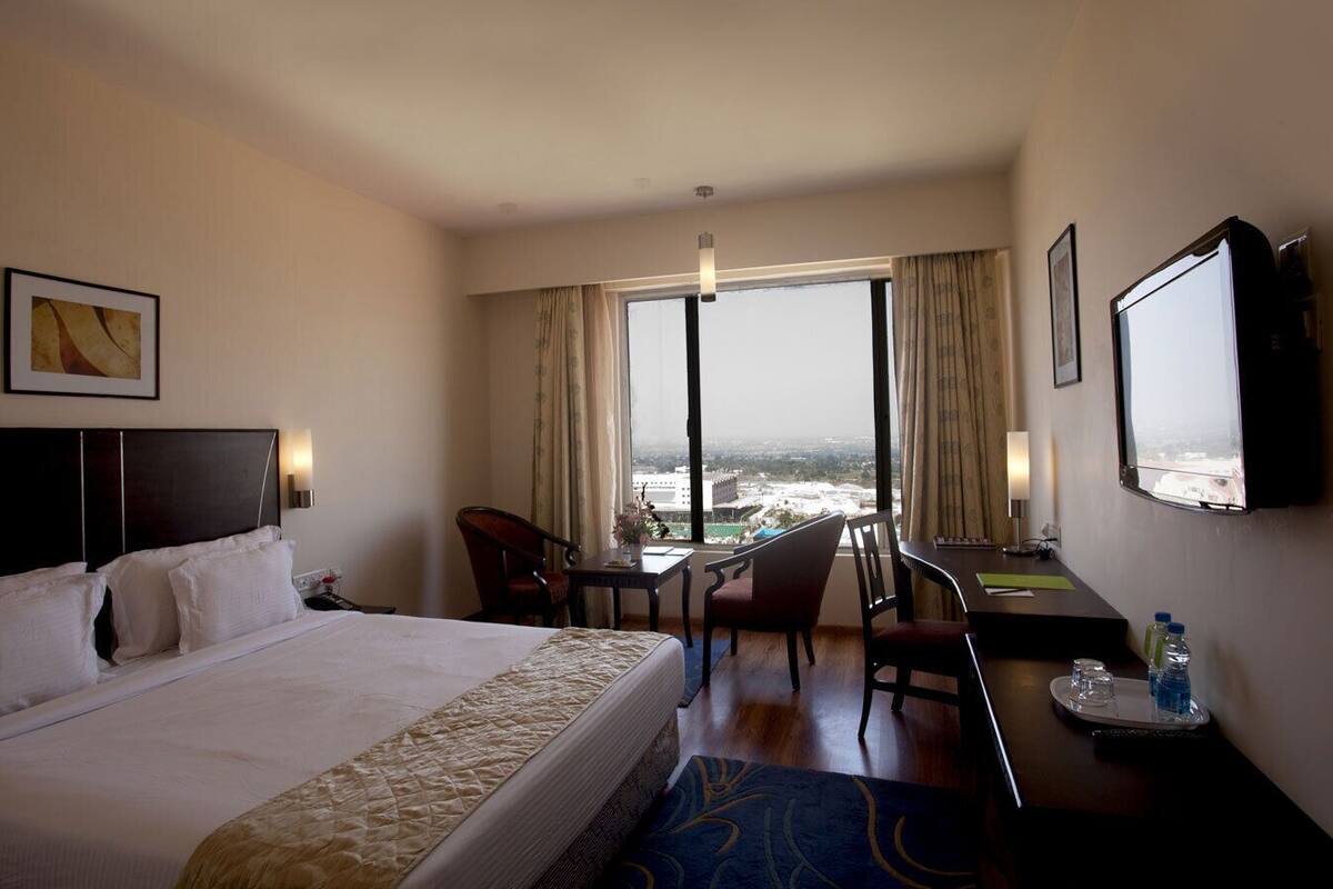 Une chambre d’hôtel avec un grand lit, un bureau, une chaise et une vue sur la ville grâce à de grandes fenêtres.